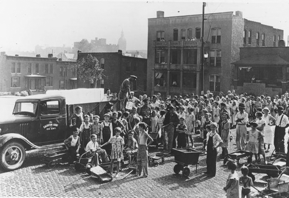 A black and white archival photo Showing an ice truck pulled up into a cobblestone street, surrounded by perhaps 100 people gathered around to get ice some with wheelbarrows. Many appear to be children, most are wearing overalls and long sleeve shirts, and the adults all have long sleeve shirts and pants and hats