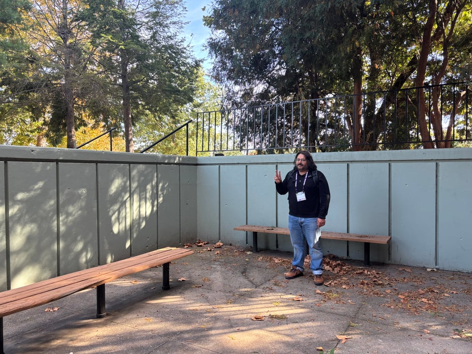 Adam standing in a sunken area outside of a dorm. There are two benches and a grey concrete wall. He gives a peace sign.