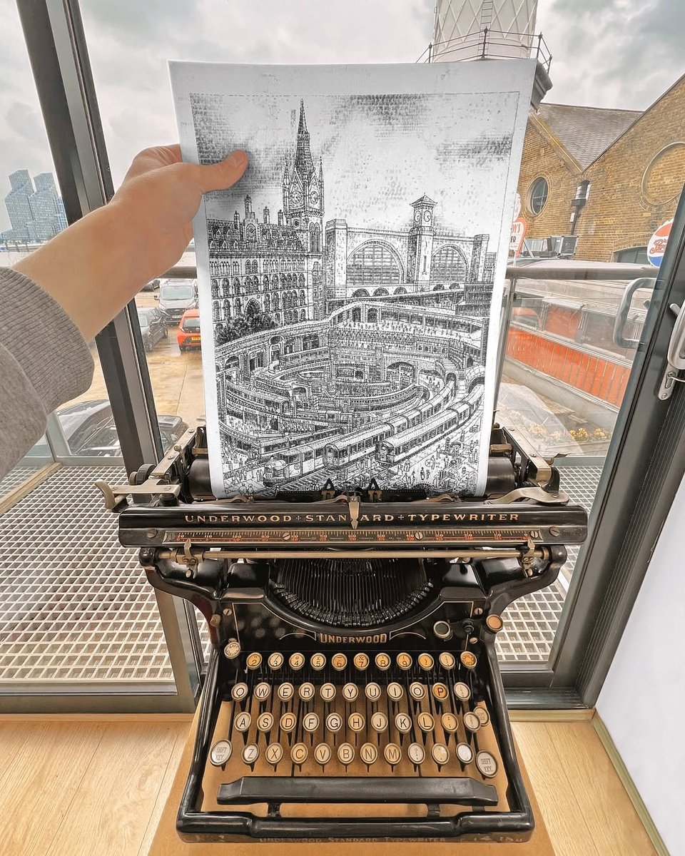 A hand holds up a detailed black-and-white drawing emerging from an antique Underwood Standard typewriter. The artwork is done entirely using thousands of individually typed characters -- and it depicts a grand railway station with arched roofs, clock towers, and multiple trains. In behind the typewriter is a modern glass-walled room overlooking city buildings and parked cars outside.