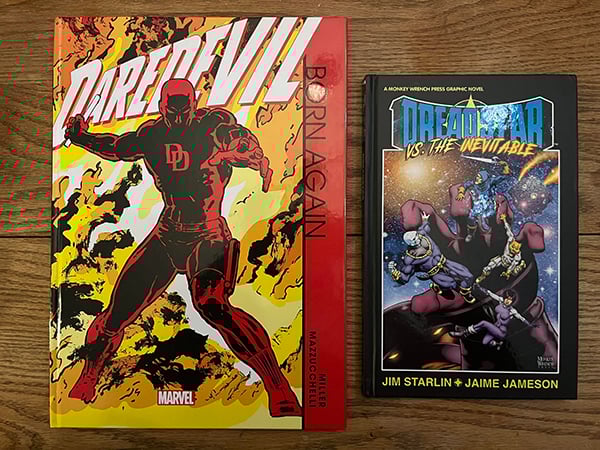 Two hardcover graphic novels sitting face-up on a table. On the left is Daredevil: Born Again, featuring a striking image of Daredevil on the cover standing in front of a fire. On the right is Dreadstar Vs. The Inevitable, featuring a band of space heroes being grabbed by the giant hand of a mysterious figure.
