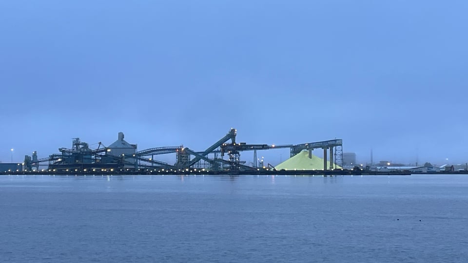 In blue-grey pre-dawn light, a small industrial complex on the shore of a body of water, consisting of conveyors and gantries around a large pile of yellow sulphur.