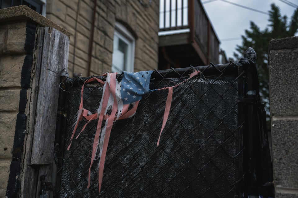 A shredded US flag hangs from a fence next to a building. The stripes hang as individual strands, and the stars section is largely intact where the flag is attached to the top of the fence.