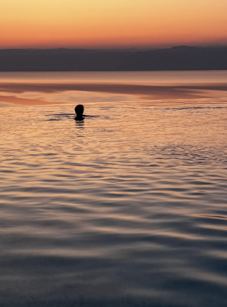 A person's head  in shadow above water and sky, all glowing orange from the recent sunset