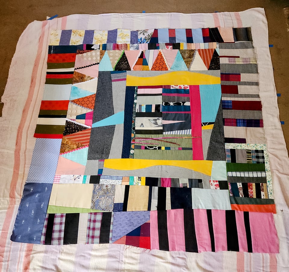 Top down photo of an improvised patchwork quilt laid on the floor for basting. The quilt has fabric in many different shapes and colors. An upcycled hospital blanket is visible around the edges of the patchwork.