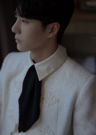 Yibo in a gorgeous white brocaded Chanel top with a short, wide, black, schoolboy-style tie. The lighting is unnecessarily dim but gives him a somber look, in contrast to the outfit, which makes him look like The Little Prince.
