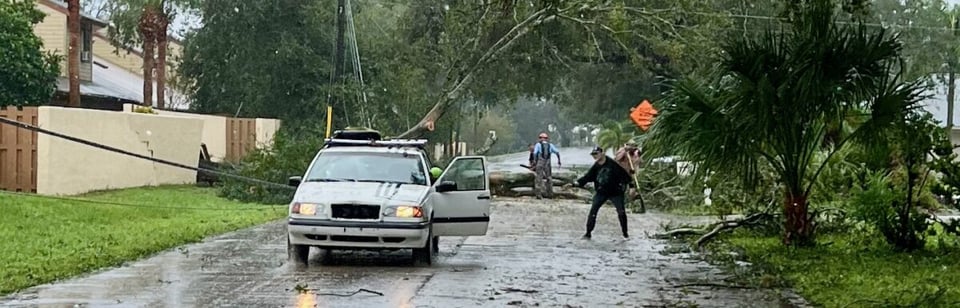 Small residential street with a large tree down across it.  To the left, you see a power line tangled in the tree.  I white Volvo station wagon is backing up to one of the freshly cut tree segments to tow it away.  Other people are gathered around the tree cutting or clearing debris to open up the roadway.