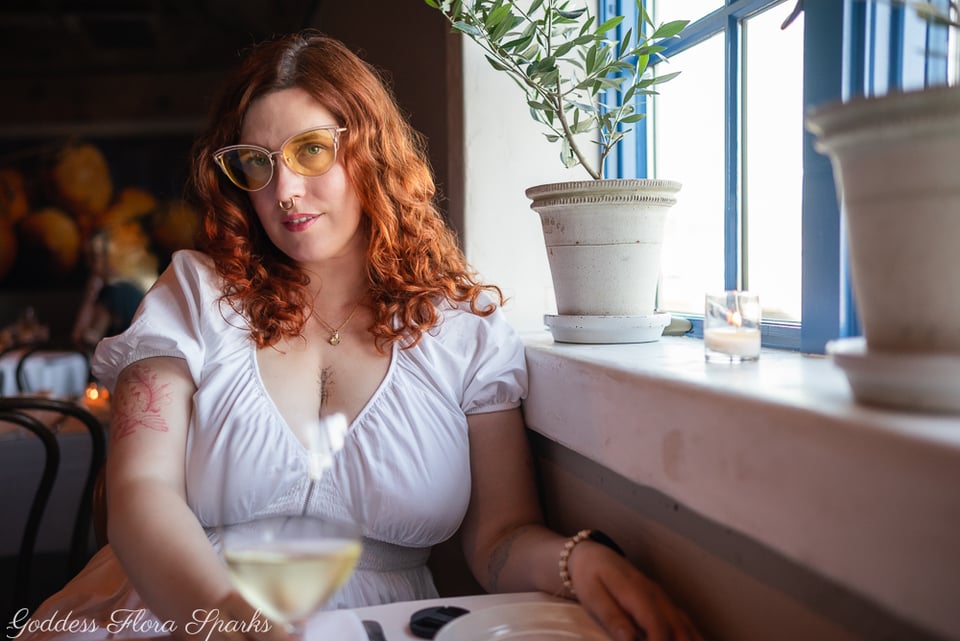 woman with red hair and white dress sits at a window lit table drinking a cool glass of sauvignon blanc