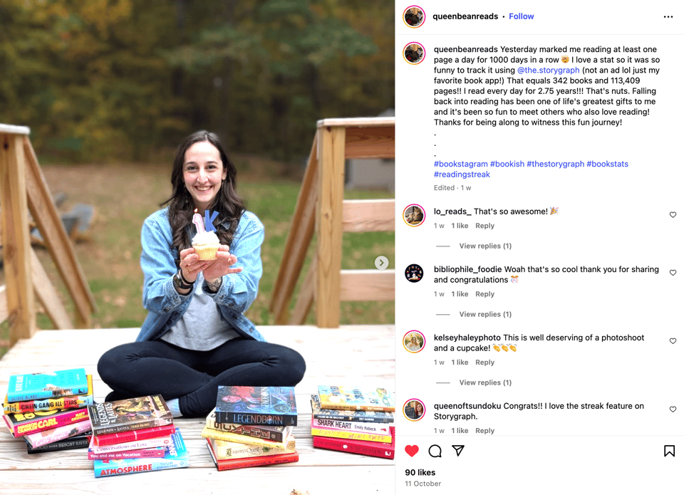 A woman sits cross-legged on a wooden deck outdoors, smiling and holding a cupcake topped with a candle shaped like the number “1K.” She is wearing a light denim shirt over a grey top and black leggings. On either side of her are stacks of colorful books arranged in piles on the deck. Behind her, the background shows trees with green and golden leaves, suggesting autumn. The Instagram caption beside the photo celebrates her reading at least one page a day for 1,000 consecutive days, tracked using The StoryGraph. She mentions reading 342 books and 113,409 pages over 2.75 years and expresses joy about rediscovering her love of reading and connecting with other readers.