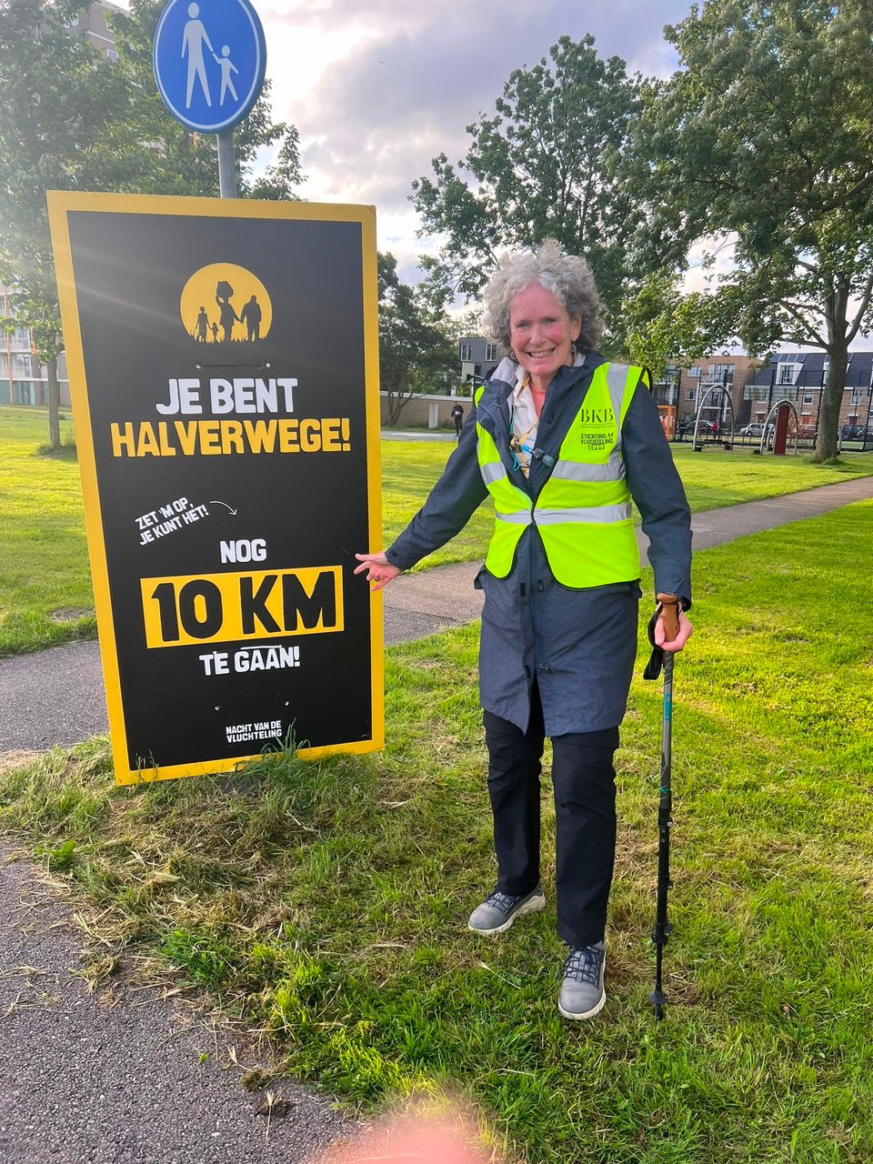 a white woman in a yellow vest and hold a walking pole stands from a sign proclaiming in Dutch that she is halfway through with 10km to go