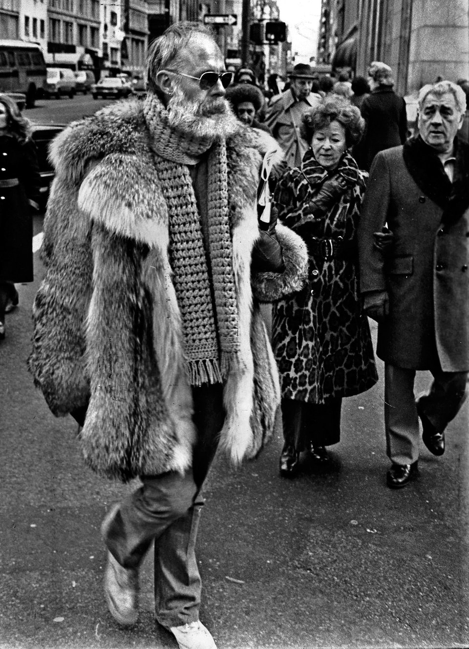 Edward Gorey wearing a fur coat, crocheted scarf, and sunglasses, stunning passersby in the street.