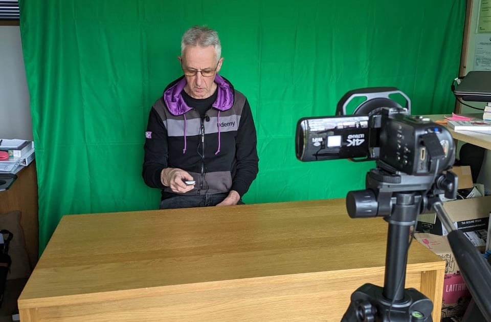 Dad sitting behind a desk, in front of a green screen, wearing a Udemy branded hoodie. There's a camera on a tripod in the foreground pointing at him. Off to the sides of the green screen are office desks and shelves.
