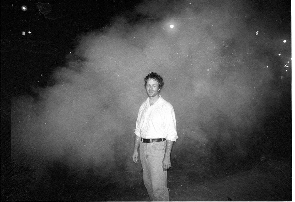 A black and white image of a person wearing light jeans and a white buttondown shirt with the sleeves rolled up, standing surrounded by a cloud of fog.