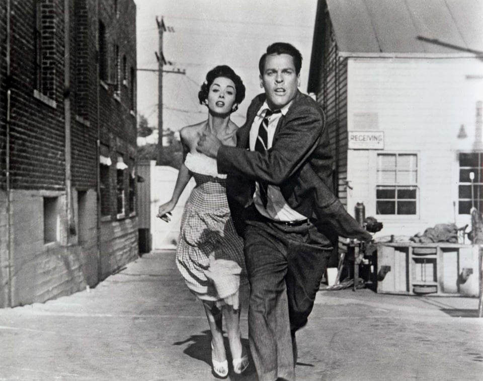 Photo still from the film, Invasion of the Body Snatchers (1956). A white man and woman flee down an alleyway in fear. They're dressed in typical 1950's middle-class attire: the man is in a dark suit and tie, the woman is wearing a light dress.