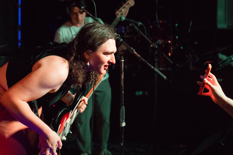 The lead singer of Vivian (left) leans forward at the edge of the stage to look at a phone that a hand holds up on the right edge of the photo