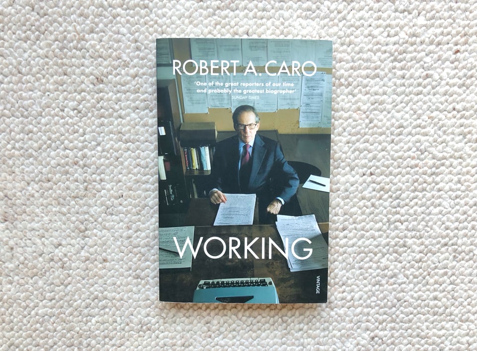 Photograph of the book, "Working", by Robert A. Caro. The cover is a photograph of the author in his office. He is wearing a suit and tie and eyeglasses. He is staring intensely into the camera. There are a typewriter and various notes on the desk in front of him. There are more pages of notes pinned to the wall behind him.nd