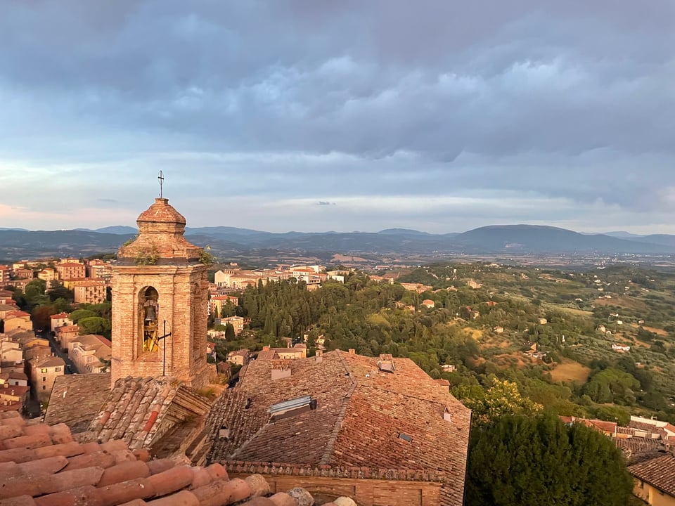 A view looking east of the Cappella di San Severo in Perugia. You can see the building's tiled roof, a stately little cupola with little saplings growing out of it, and in the distance the green countryside of Umbria with the Apennine Mountains in the distance