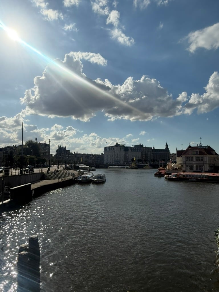 A canal scene in Amsterdam under a bright blue sky with large white clouds. Sunlight beams dramatically through a cloud, casting rays across the sky. Several boats are docked along the water, including sightseeing boats. On the left, stone steps and a quay with people walking lead into the view. In the background, historic Amsterdam buildings line the waterfront, their outlines softened by the sunlight. The water in the foreground sparkles with reflections of the sun.