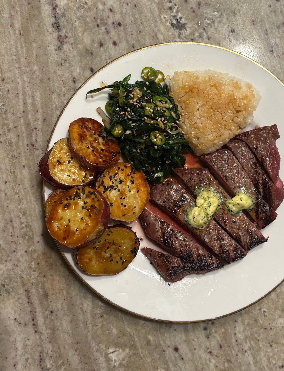 A plate of sliced steak topped with shio koji-infused butter, browned satsuma potatoes with black sesame seeds, sautéed greens, and toasted onigiri.