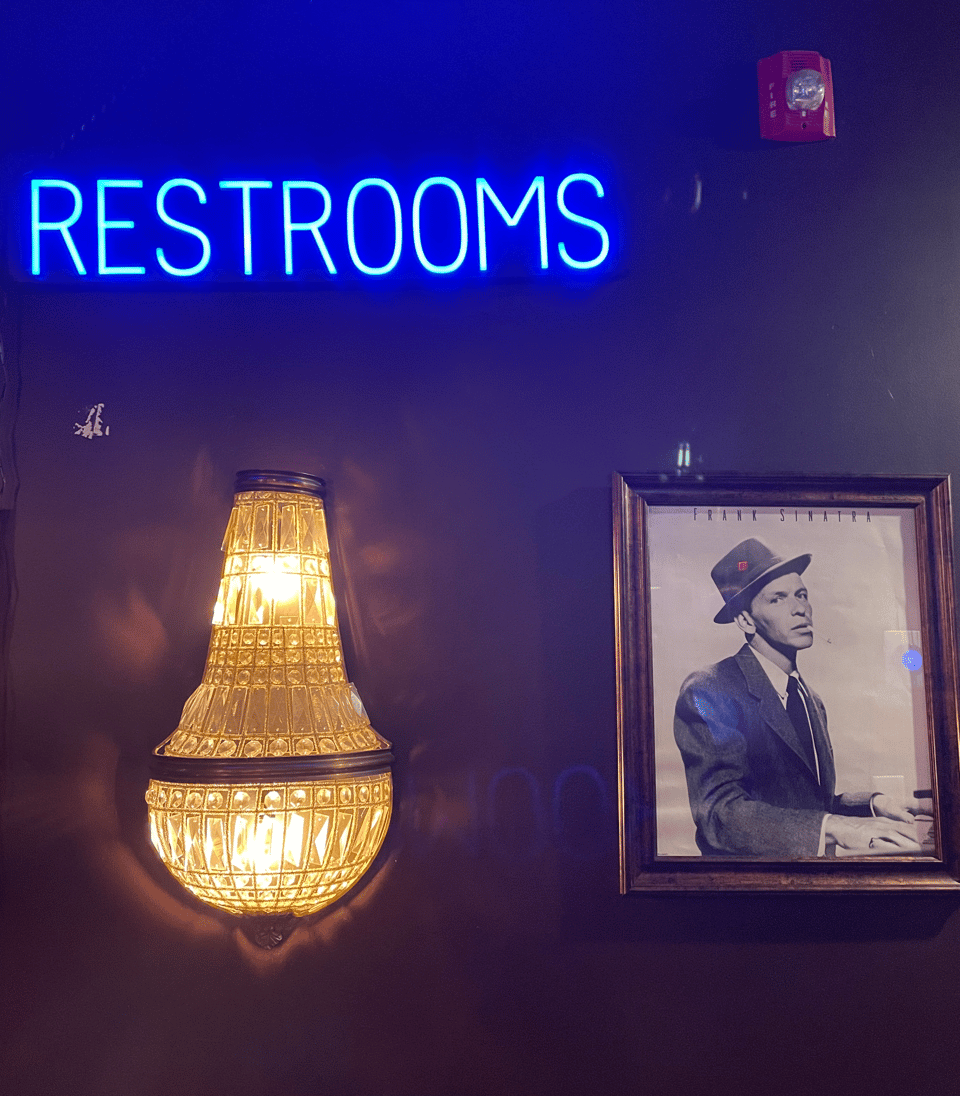 An elegant wall with a fancy wall light, a blue neon sign that says RESTROOMS and a promo pic of mid 1950s Frank Sinatra glaring at the viewer