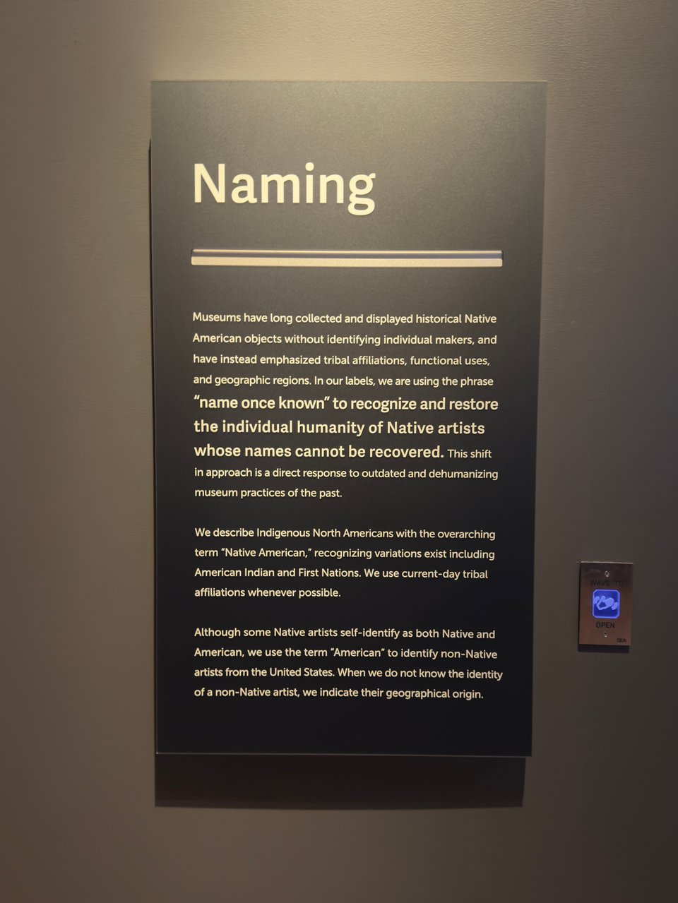 Sign on brown wall that says: Museums have long collected and displayed historical Native American objects without identifying individual makers, and have instead emphasized tribal affiliations, functional uses, and geographic regions. In our labels, we are using the phrase
"name once known" to recognize and restore the individual humanity of Native artists whose names cannot be recovered. This shift in approach is a direct response to outdated and dehumanizing museum practices of the past.
We describe Indigenous North Americans with the overarching term "Native American," recognizing variations exist including American Indian and First Nations. We use current-day tribal affiliations whenever possible.
Although some Native artists self-identify as both Native and American, we use the term "American" to identify non-Native artists from the United States. When we do not know the identity of a non-Native artist, we indicate their geographical origin.
