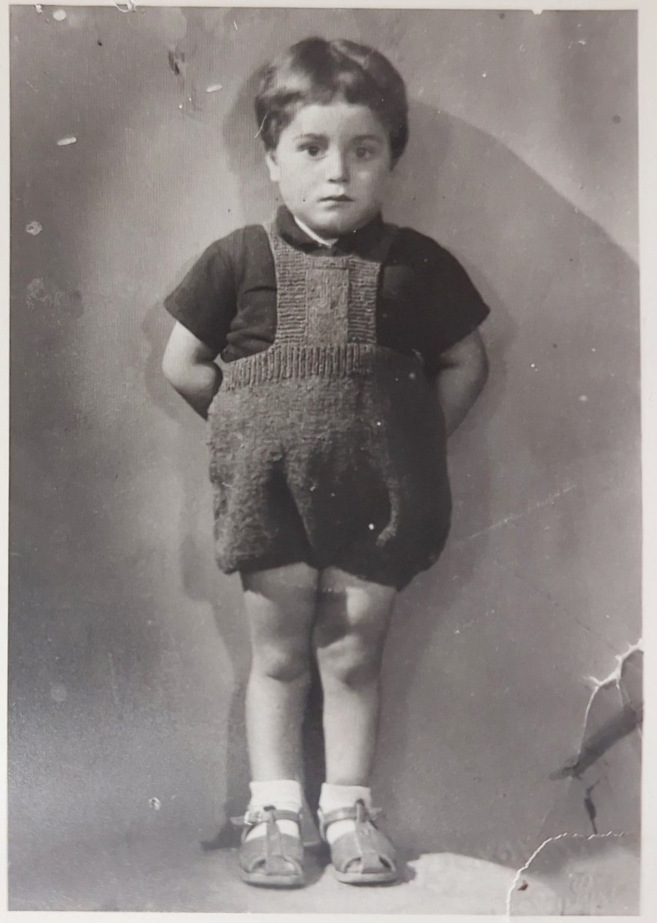 A black and white photo of a little boy in a knitted onesie, wearing socks and sandals, standing against a wall looking afraid. It was his first formal picture and he told me was very scared.