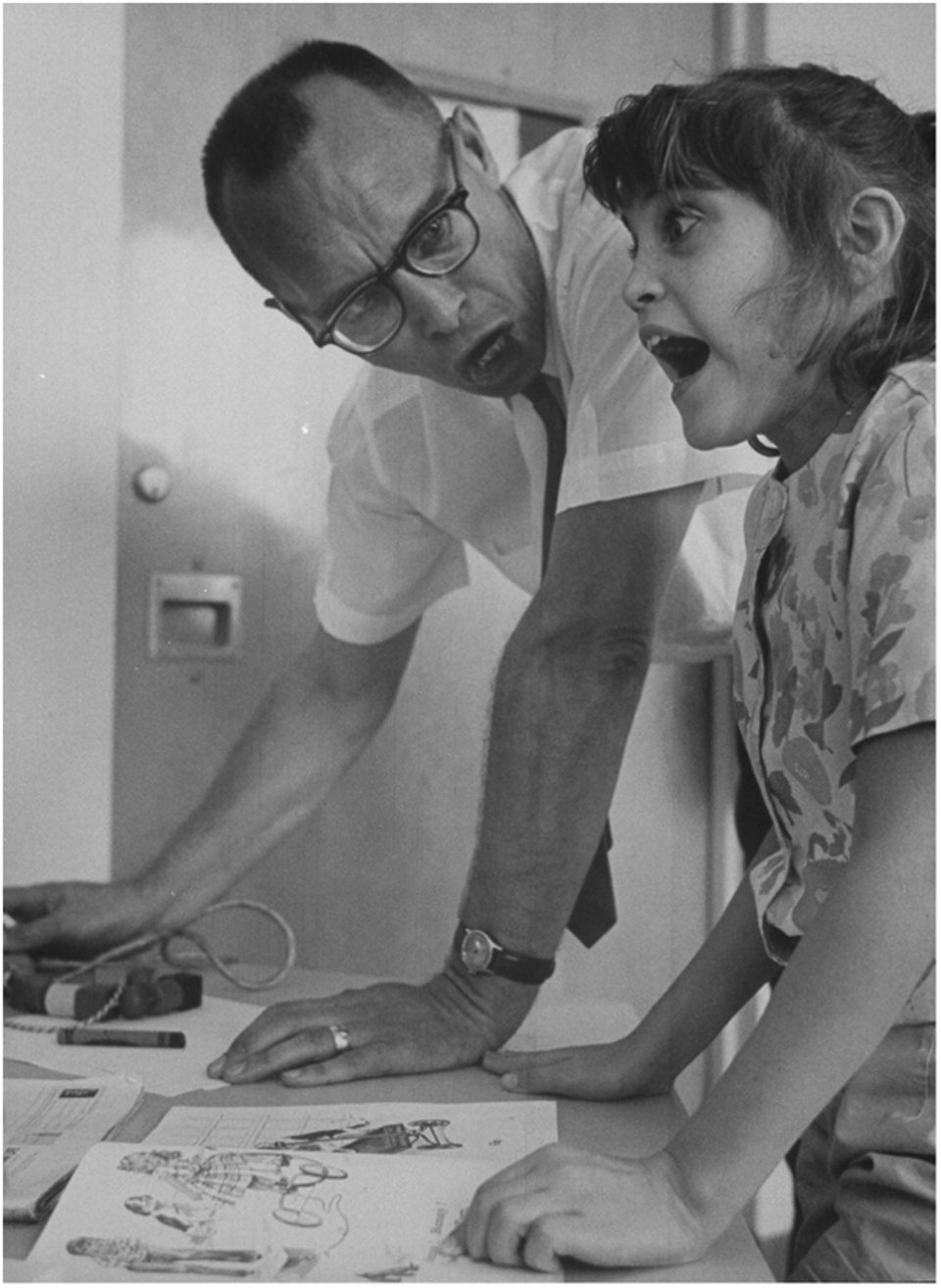 A white man resting his hand on a desk next to a white child. He is wearing glasses, a short sleeved buttondown, a tie, and a watch, and is making a theatrical frowning expression as he looks at her. She has brown hair, bangs and a ponytail, wearing a flowered buttoned shirt, also resting her hands on the desk, is looking straight ahead with her mouth open. Black and white photo.
