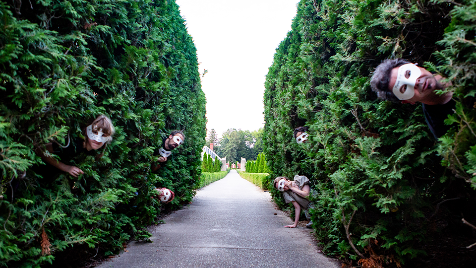 Faces in masks peer out from the foliage of a hedge maze. It is unsettling, but not creepy.