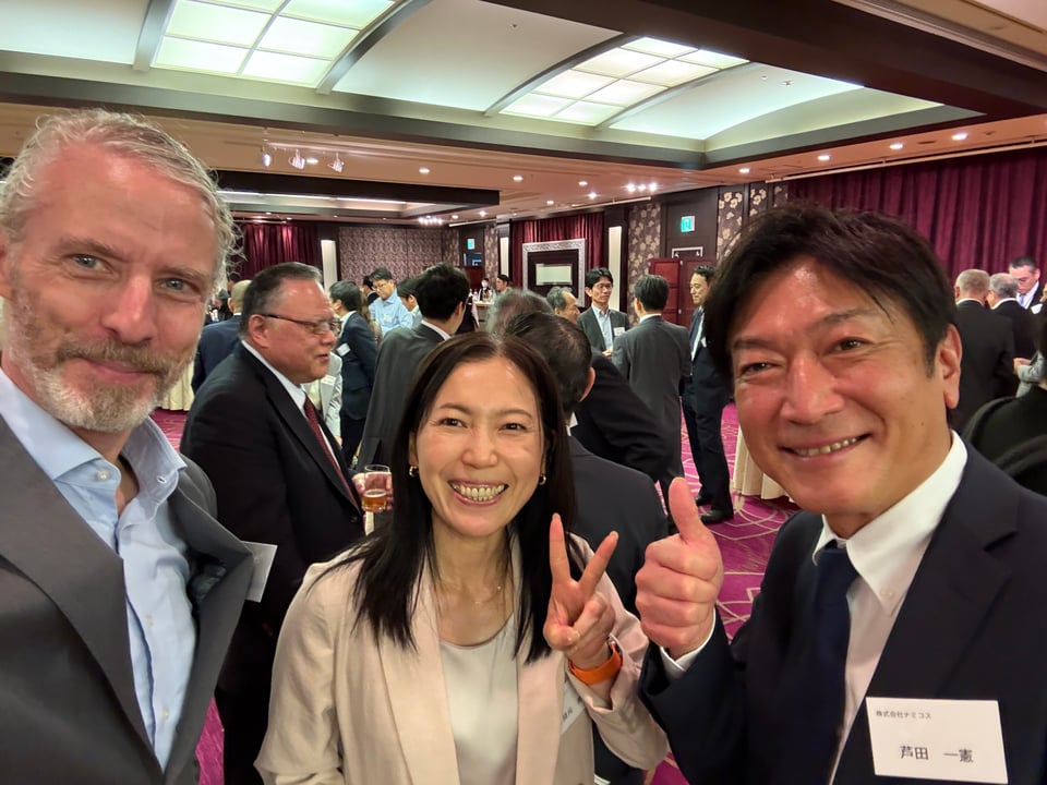 Selfie of a white man, Japanese woman and Japanese man at a business reception. The woman is holding up a peace-V, and the Japanese man is giving a thumbs-up. They all look happy.