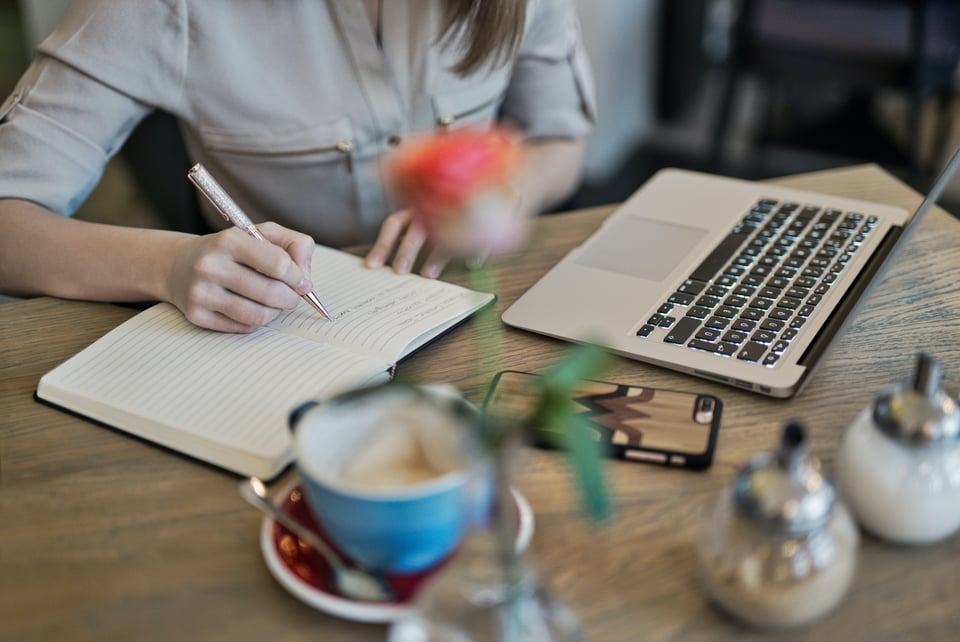 The image of a person (no face shown) writing in a notebook at a table. There is also a cellphone, a cup of tea, and an open laptop on the table. There is also a small vase with a rose on the table, but it's blurred out to focus specifically on the person, notebook, and laptop.