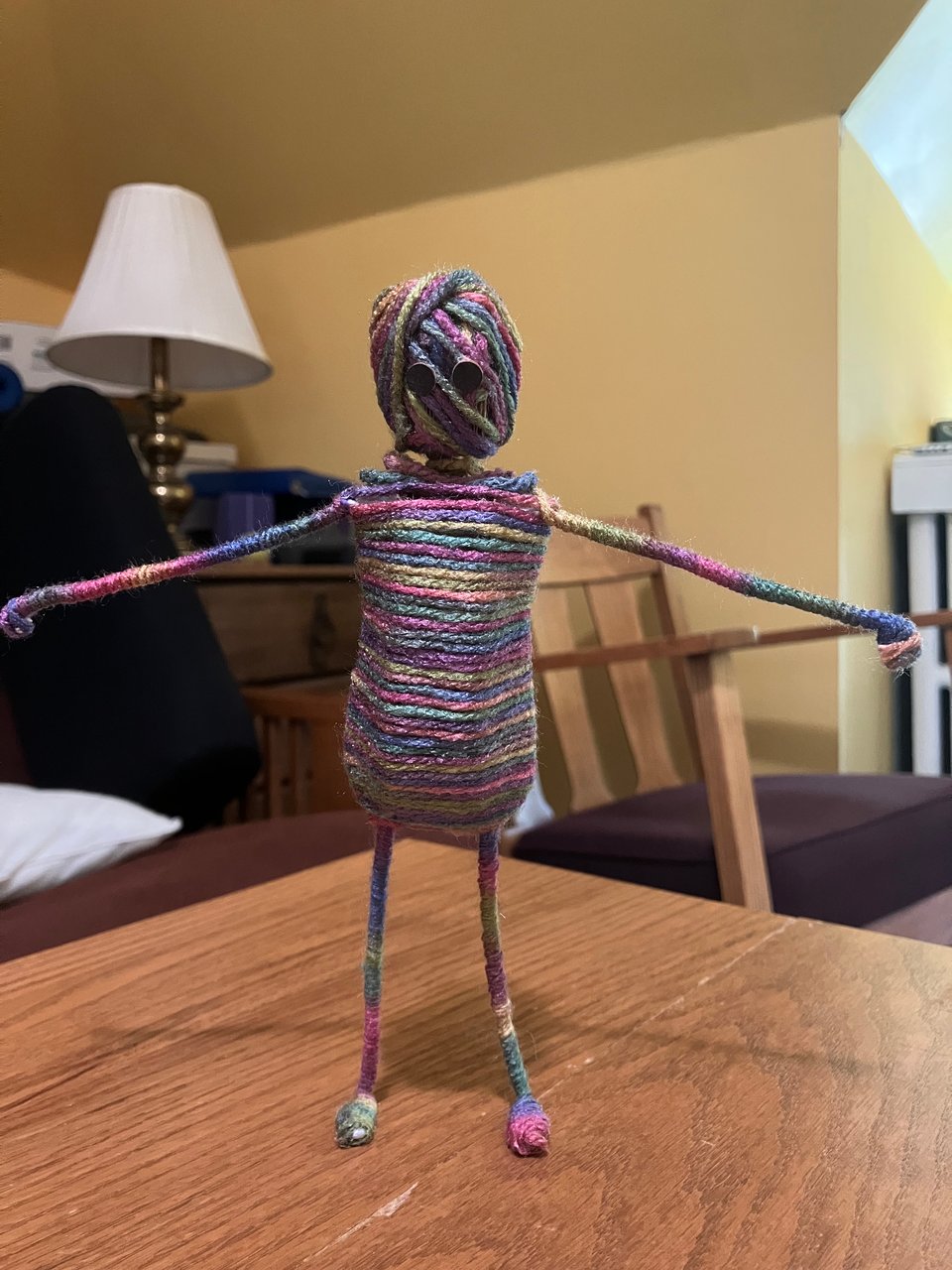 A humanoid puppet, wrapped in multicolored yarn, with skinny limbs and large black eyes, stands on a table and looks straight at the camera, with arms outstretched to his sides. Behind him, irrelevantly, is a couch, lamp, and rocking chair.