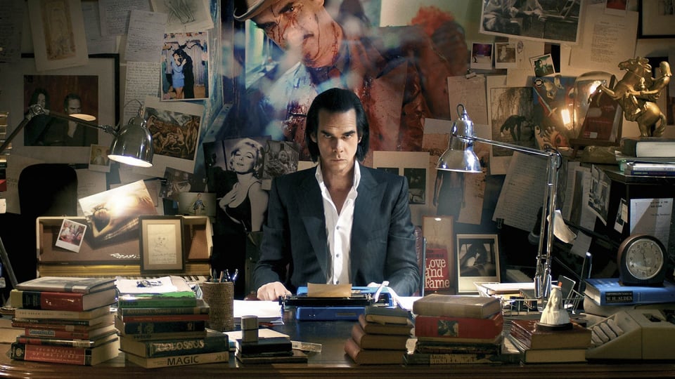 photo from the movie 20,000 DAYS ON EARTH, showing the musician Nick Cave sitting at a desk with typewriter, surrounded by books and with a wall of photos and images behind him