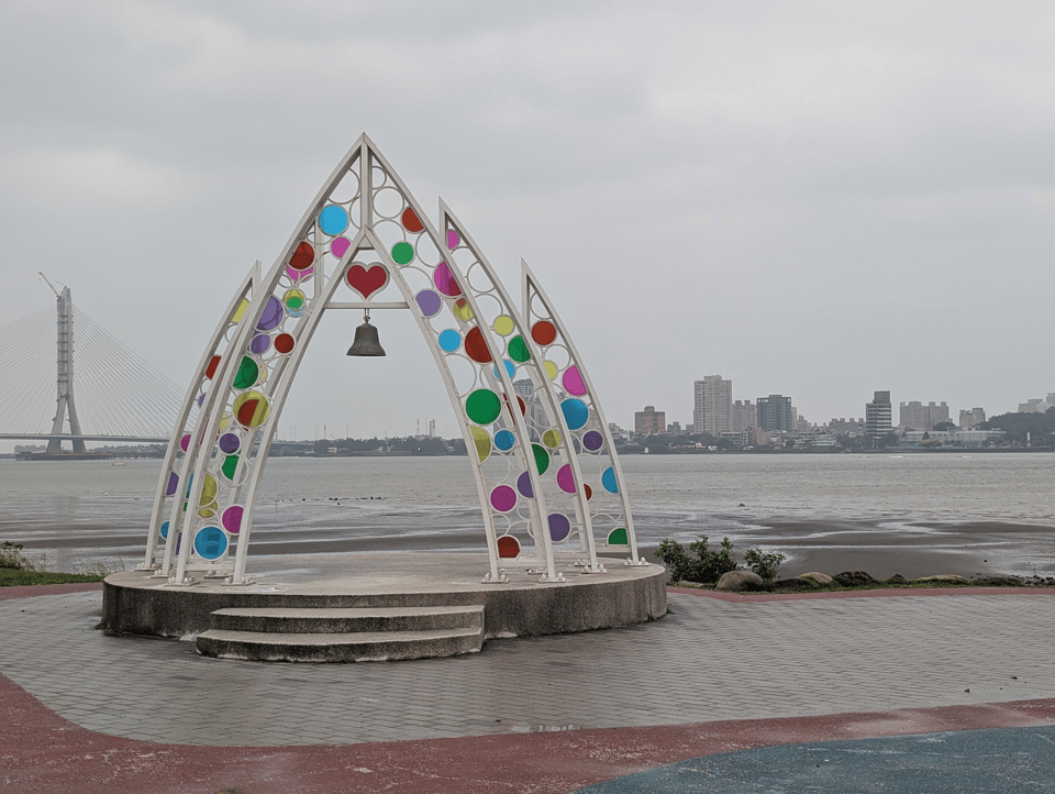 A riverside platform with a colorful arch above it, reminiscent of a miniature wedding altar. The arch has many colorful circles of stained plexiglass, but the background is very grey and dreary. Across the water you can see an urban skyline.