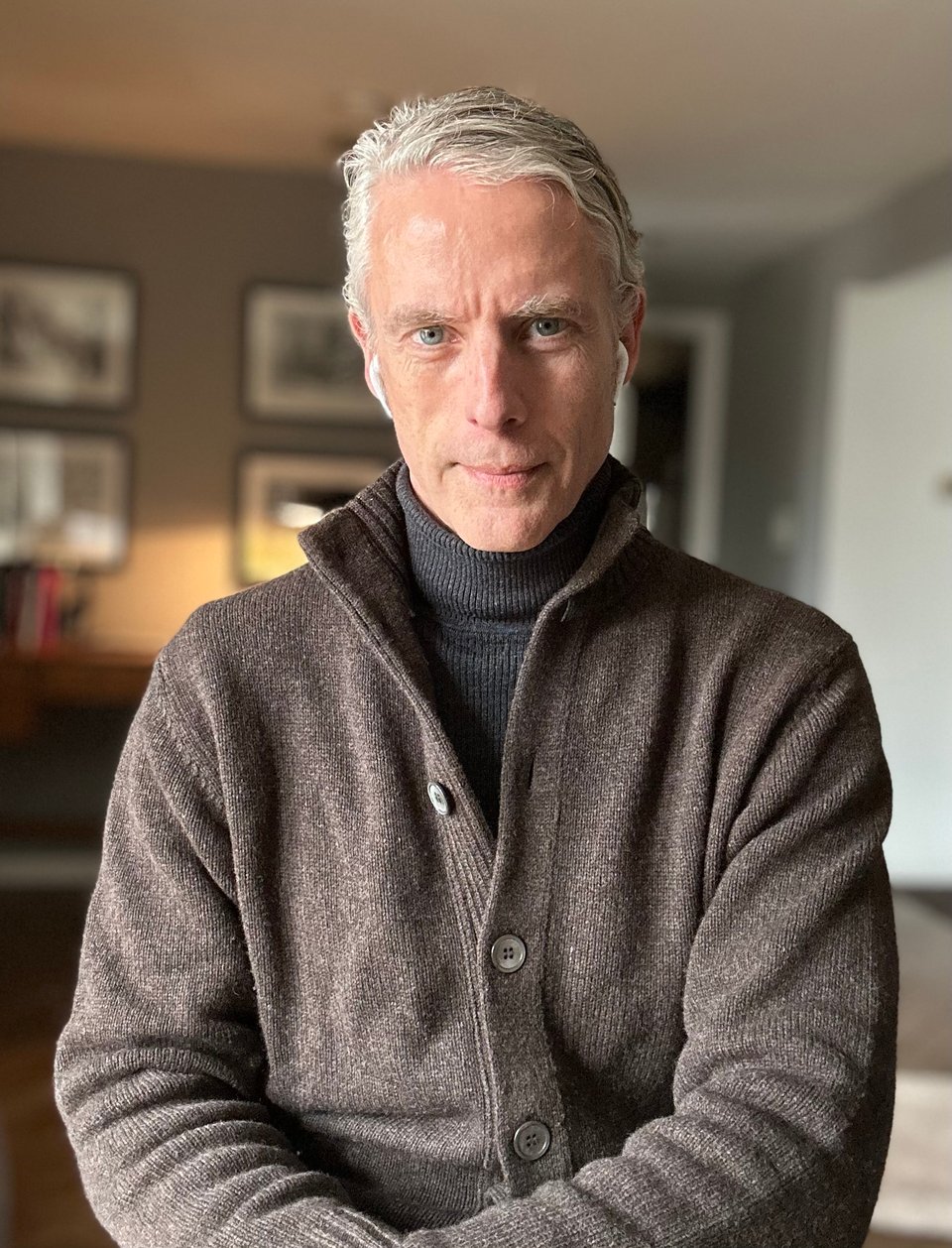 Portrait-setting iPhone photo of a white man in a dark brown cardigan over a dark gray ribbed turtleneck. He has a pair of early-style AirPods in, and the left side of his face is partly in shadow. He looks alright; at least, you wouldn't notice that he's absolutely walloped by a head cold.