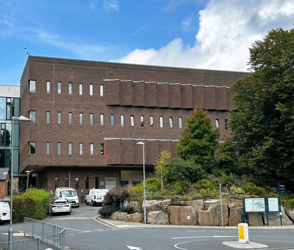 A four storey dark red brick building. The upper storeys overhang the ground floor where there are clearly services at work based on all the vans. The lines of narrow slit windows are disrupted by the triangular bays which, from this angle, present blank brick walls.