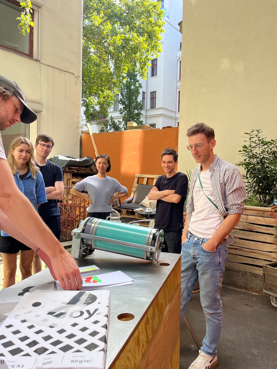 Photo of a group of people standing outside around a table, looking at some printed material and a paint roll for a riso printer