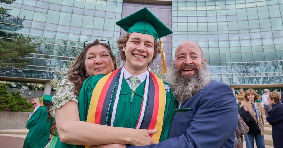 JMC graduate wearing a green cap and gown, embraced on either side by smiling family members outside of the Wharton Center.