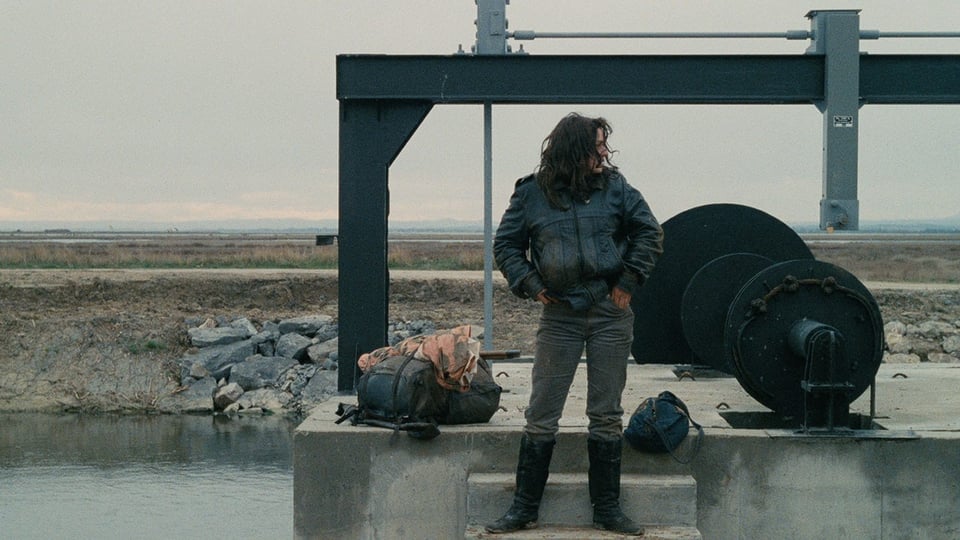 Vagabond protagonist Mona in winter wear standing in front of water well equipment in the French winter countryside.