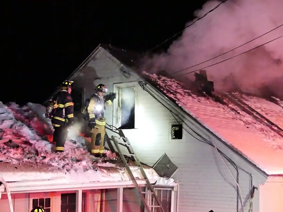 Firefighters standing outside a house with lights on. They are wearing full firefighting gear including helmets, boots, gloves, and protective clothing. The house has visible damage to the roof and siding.