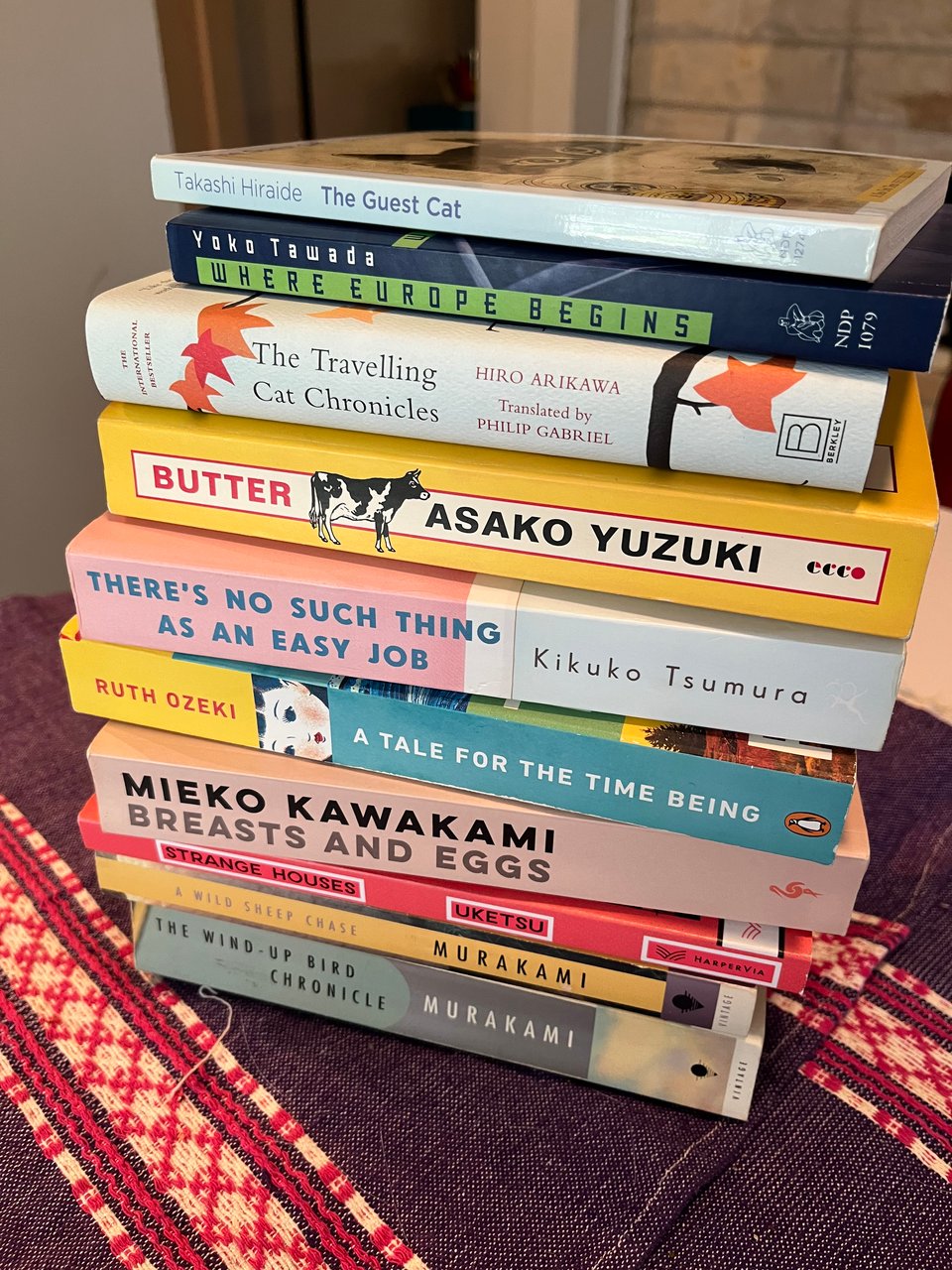 A stack of books by Japanese and Japanese-American authors, all published in English, sit on top of a table covered with blue and pink place mats.