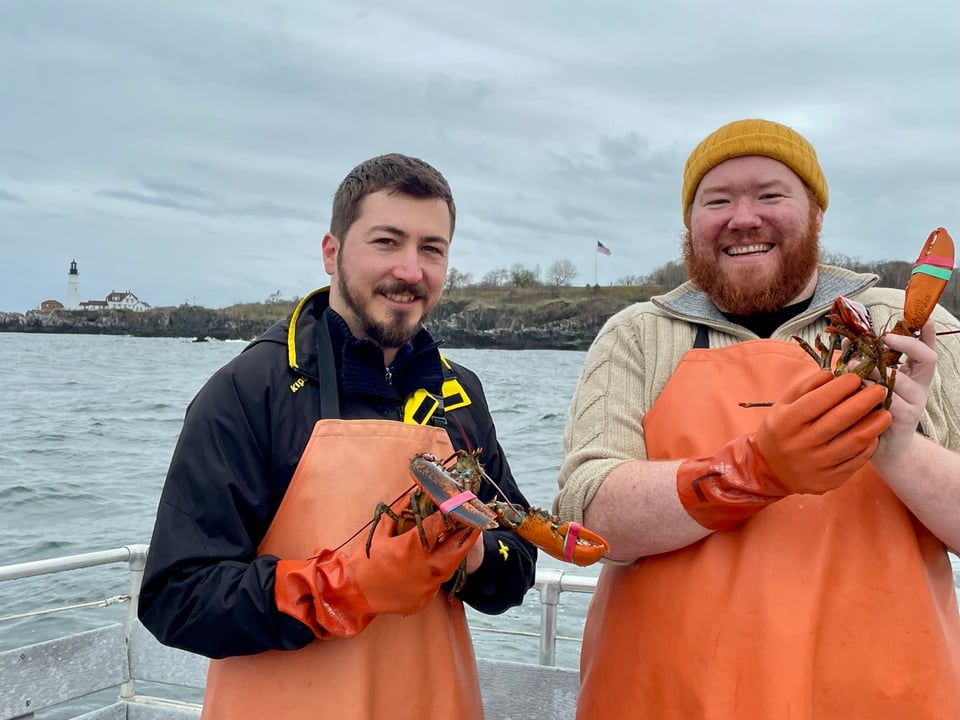 A photograph of me, Victor, and my partner Henry on a boat, both wearing orange aprons and holding lobsters in our hands