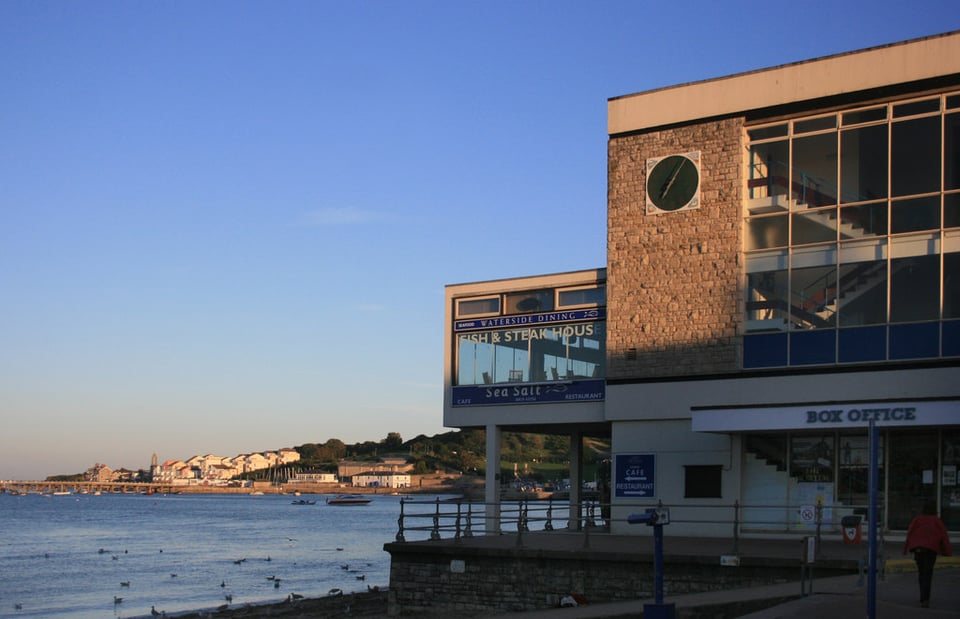 A 1960s brick and glass building on a seafront, caught in evening sunlight. The main block is three storeys high. On the seaward side, a glass single storey enclosed balcony juts out over pilotti. Signage offers fish and steaks.