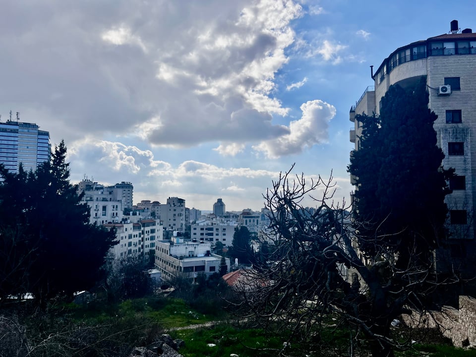 A view from a hilltop of a small city of off-white buildings,  blue sky with clouds, over a small city of off-white buildings