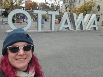 Meredith wears a hat and scarf and poses in front of a sign that says OTTAWA.