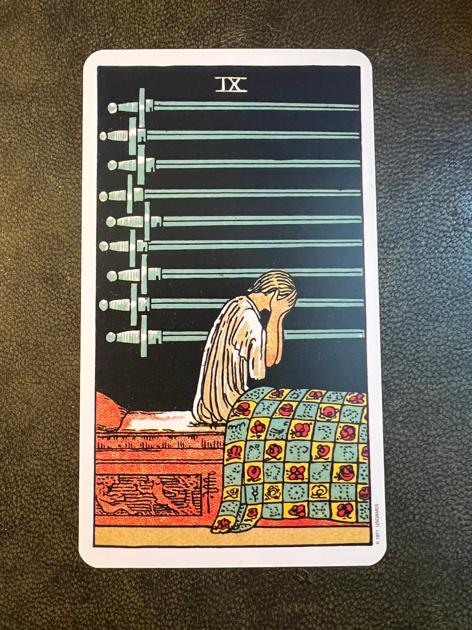 A woman in side profile, sitting up in bed with her hands over her face. There are nine swords on a black wall behind her, all pointing in the direction she's facing.