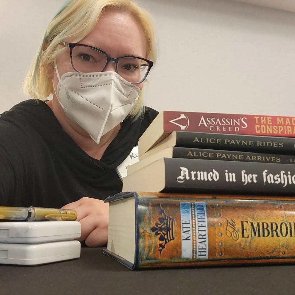 Kate is wearing glasses and a mask and sitting behind a stack that contains the Alice Payne novellas, The Embroidered Book, Armed in Her Fashion, the first Assassin's Creed book. The little white containers are ink for stamps, and there's a gold ballpoint pen on top.