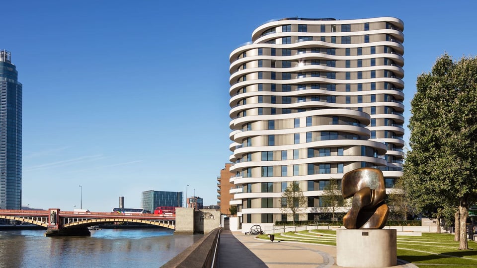 A landscape view of the building, with the Thames to the left. Tan cement building with windows reflecting the sun, multiple layers of curvatures providing balcony space. Very mod upper-crust city apartment building design, if you know you know. Park with trees to the right, modern sculpture at front.