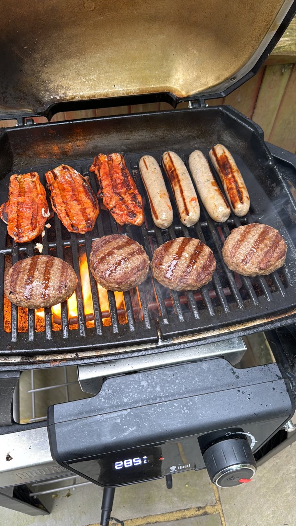 A picture of the barbecue open and cooking with burgers and sausages on it.