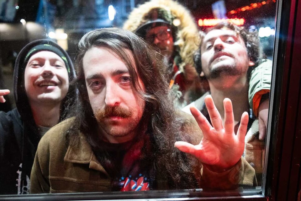Four men are shown through a window. One man, with a mustache and long hair, has his left hand pressed against the glass.