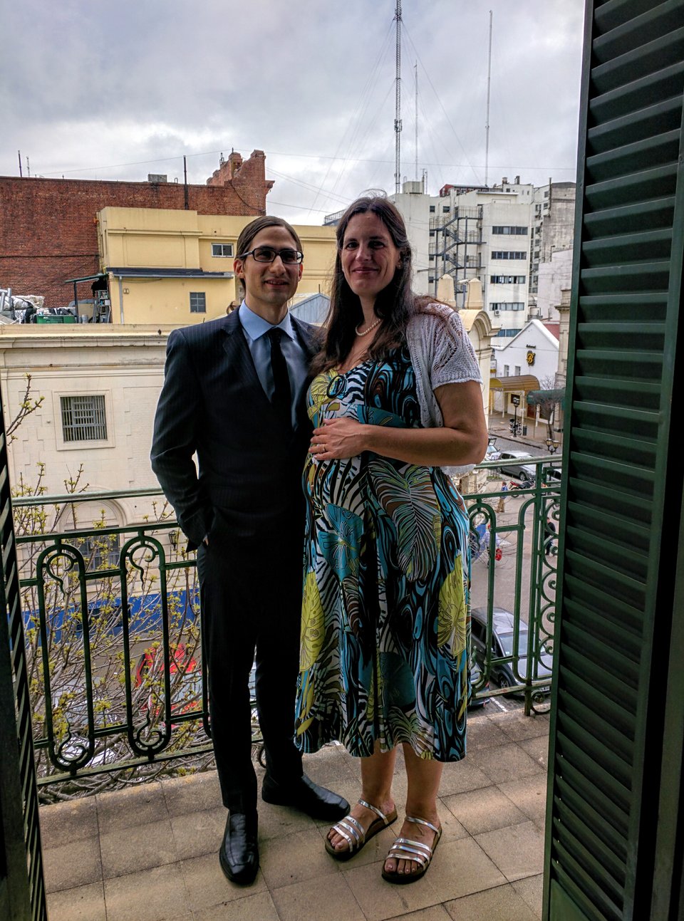 A couple dressed up for their wedding stand on a balcony. The man is wearing a navy suit and the woman wears a colorful dress and has her hand on her pregnant belly.