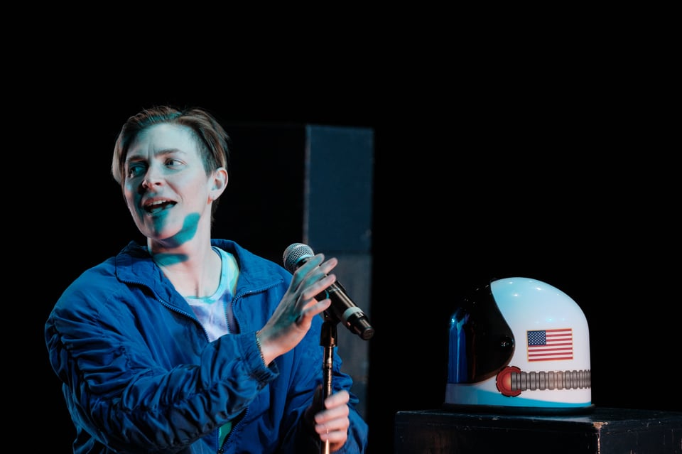 Nora looks out at the audience invitingly, with their mouth open, about to speak. they're reaching for a microphone on a stand. an astronaut helmet sits on a stack of boxes nearby them.